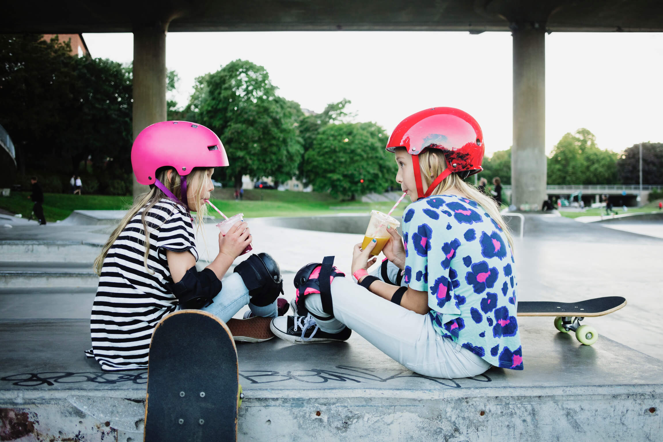 Två små tjejer med hjälm, armbågsskydd och skatebord sitter i en skatepark och dricker juice